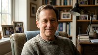Middle-aged Caucasian man with light brown hair wearing a beige knit sweater, seated in a comfortable chair in a home library setting with bookshelves and framed photos visible behind him.