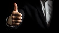 Businessman in dark suit showing thumbs up gesture against black background, conveying approval and positive feedback in professional setting.