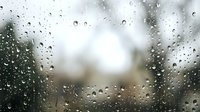 Raindrops cover glass window with blurred outdoor scenery behind, creating moody atmospheric effect suitable for weather, seasonal, and emotional design projects.