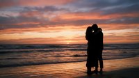 Romantic couple silhouette embracing on beach at sunset with dramatic orange and pink sky reflecting on ocean waves creating intimate moment.