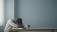 Exhausted man resting head on desk beside coffee mug and notebook, expressing burnout, stress, or fatigue in a minimalist blue room.