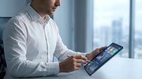 Young Caucasian businessman in white shirt using tablet with colorful analytics dashboard in modern office, city view background.