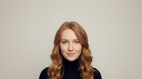 Young Caucasian woman with wavy auburn hair wearing black turtleneck sweater smiling at camera against neutral beige background for portrait photography.