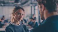 Young woman with blonde hair in casual business attire engaged in serious conversation with male colleague in modern office environment with blurred background.