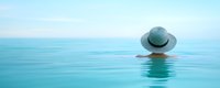 Woman in white sun hat relaxing in infinity pool with turquoise water against blue sky horizon, view from behind creating serene vacation atmosphere.