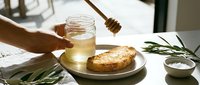 Honey drizzling from wooden dipper into glass jar above fresh bread and rosemary sprigs on table for food styling and culinary content.