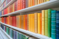 Colorful rainbow collection of vintage books arranged by color on white library shelves, creating ombre gradient effect from red through yellow to green and blue.