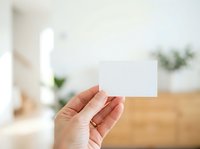 Caucasian hand holding blank white business card in modern home office environment with wooden desk and blurred green plants in background for branding mockup.