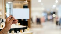 Hand holding blank white card in retail store environment with blurred shopping mall background, suitable for promotional messaging and marketing campaigns.