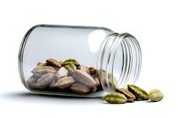 Roasted pistachios spilling from clear glass jar on white background, nuts scattered around container showing natural green and brown colors.