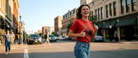 Young Hispanic woman laughing joyfully while walking down sunny urban street with historic buildings and shops in background during golden hour.