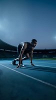 Athletic African American male sprinter in starting position on blue running track at stadium during evening training session for competitive sports marketing.