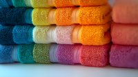 Colorful stacked bath towels in rainbow gradient from blue to pink, showing soft cotton terry texture and decorative borders, arranged in neat rows.