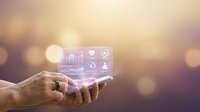 Business professional holding smartphone with holographic digital interface displaying analytics charts and icons against warm bokeh background for technology presentations.