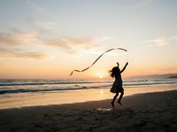 Silhouetted female child playing with ribbon on sandy beach at golden hour sunset, ocean waves in background, outdoor recreation and leisure activity.