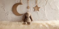 Soft plush bunny toy sitting on fluffy white fur against textured wall with hanging golden moon and stars decorations, creating cozy nursery atmosphere.