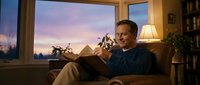 Middle-aged Caucasian man relaxing in comfortable armchair reading book at home during sunset. Cozy living room interior with warm lighting and plants.