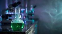 Green chemical solution in round bottom flask on laboratory table with blurred scientific equipment and colorful lighting in background for research.
