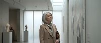 Middle-aged woman with blonde hair wearing a beige blazer stands in a modern art gallery or museum corridor, viewing contemporary artwork on white walls with natural skylight illumination.