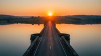Straight road stretching toward golden sunrise over calm water, symmetrical perspective with sun on horizon, ideal for travel and journey concepts.