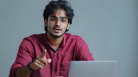 Young South Asian man with glasses in burgundy shirt pointing at camera while sitting with laptop, conveying direct communication in professional setting.