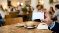 Man in business attire holding blank white card at wooden table with soup bowl, blurred office lunch meeting background with colleagues and festive decorations.