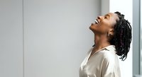 Young African American woman with braided hair wearing white shirt, head tilted back in relaxed pose against white wall. Suitable for wellness, mental health, stress relief, and lifestyle content.