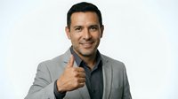 Middle-aged man with dark hair and Mediterranean ethnicity wearing gray blazer over dark shirt, giving thumbs up gesture with confident smile against white background.