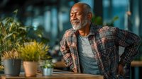 Mature African American man experiencing back pain in modern office environment, holding lower back while standing near desk with plants.