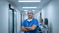 Middle-aged Caucasian male surgeon in blue scrubs and surgical cap standing confidently in modern hospital corridor with arms crossed.