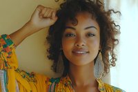 Young African American woman with natural curly hair wearing yellow floral dress and statement earrings smiling confidently against light background, expressing joy and authenticity.