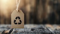 Recycling symbol on kraft paper tag with string against blurred natural background, promoting environmental awareness and sustainable lifestyle choices.