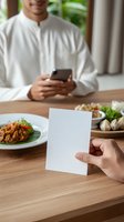 Asian man in traditional white clothing holding smartphone at dining table with local cuisine, while hand holds blank white card in foreground.
