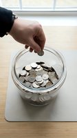 Hand dropping a coin into a glass jar filled with silver coins on a wooden table, symbolizing savings, budgeting, and financial growth.