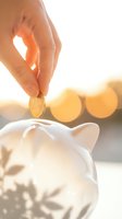 Hand dropping golden coin into white piggy bank with warm sunset bokeh lights creating financial savings and investment concept for retirement planning.