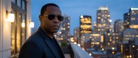 Middle-aged Black male professional wearing sunglasses and dark blazer against illuminated city skyline at dusk, urban corporate lifestyle portrait.