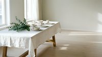 Minimalist dining table setup with neutral linen tablecloth, place settings, and eucalyptus greenery by bright window in modern interior space.