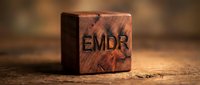 Wooden cube block with EMDR letters engraved on natural wood surface representing eye movement desensitization reprocessing therapy concept for mental health treatment.