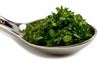 Fresh chopped parsley leaves on stainless steel serving spoon against white background, ready for cooking and food presentation.