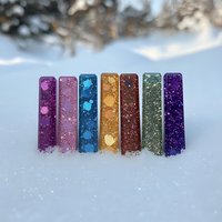 Colorful glittery resin bookmarks arranged in rainbow order standing in fresh snow, creating magical winter atmosphere with sparkles and bokeh effect in background.
