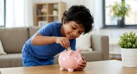 Young Hispanic boy in blue shirt putting coin into pink piggy bank at home teaching children financial literacy and saving money habits.