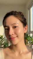 Young Asian woman with natural glowing skin and messy bun hairstyle smiling at camera in bright indoor setting with plants for skincare and beauty content.