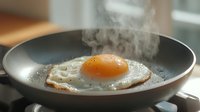 Steaming sunny side up egg frying in black pan with rising smoke, bright orange yolk and crispy white edges, morning breakfast cooking process.