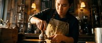 Young Asian male barista in apron tamping coffee grounds in portafilter at rustic cafe, warm moody lighting, craft espresso preparation.