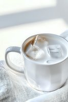 White ceramic mug filled with creamy drink and ice cubes on light linen fabric background, soft natural lighting creates gentle shadows and highlights.