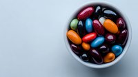 Colorful jelly beans in white ceramic bowl on light background, overhead view showing vibrant candy assortment with glossy surface and mixed flavors.