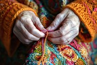 Senior woman's wrinkled hands in orange sweater knitting colorful wool yarn. Close-up view shows detailed texture of handmade craft project with vibrant pattern.