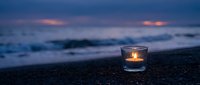 Glowing candle in glass holder on pebble beach at twilight with ocean waves and purple sky creating peaceful meditation atmosphere for relaxation concepts.