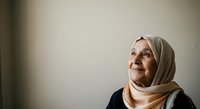 Senior woman wearing beige headscarf and dark clothing, looking upward with contemplative expression against neutral background. Suitable for healthcare, wellness, spirituality, and diversity concepts.
