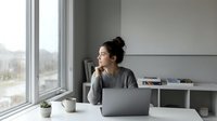 Young Asian woman working remotely from home office, sitting at desk with laptop computer, drinking coffee while looking out window during daytime work session.